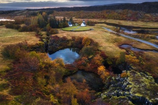 A white church sits on a grassy plain surrounded by autumn-colored trees and streams in Thingvellir National Park.