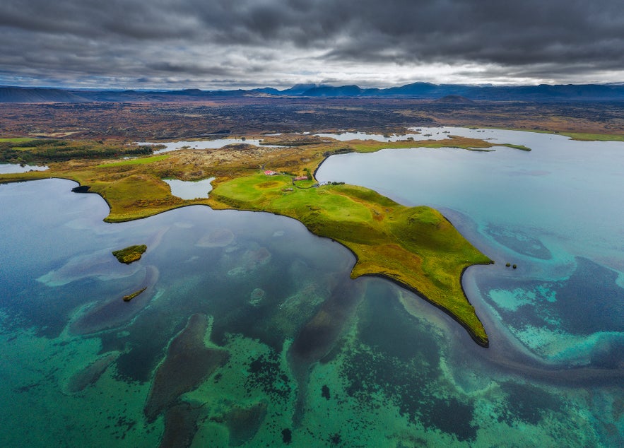 Vista panoramica aerea del Lago Myvatn, un’altra attrazione top in Islanda, con acque turchesi che rivelano formazioni laviche sotto la superficie. Una penisola verde si protende nel lago, incorniciata da campi e paesaggi vulcanici sotto un cielo spettacolare
