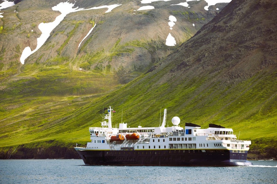 Passenger ferry to Iceland sailing by steep green mountains with patches of snow in a scenic East Iceland fjord.