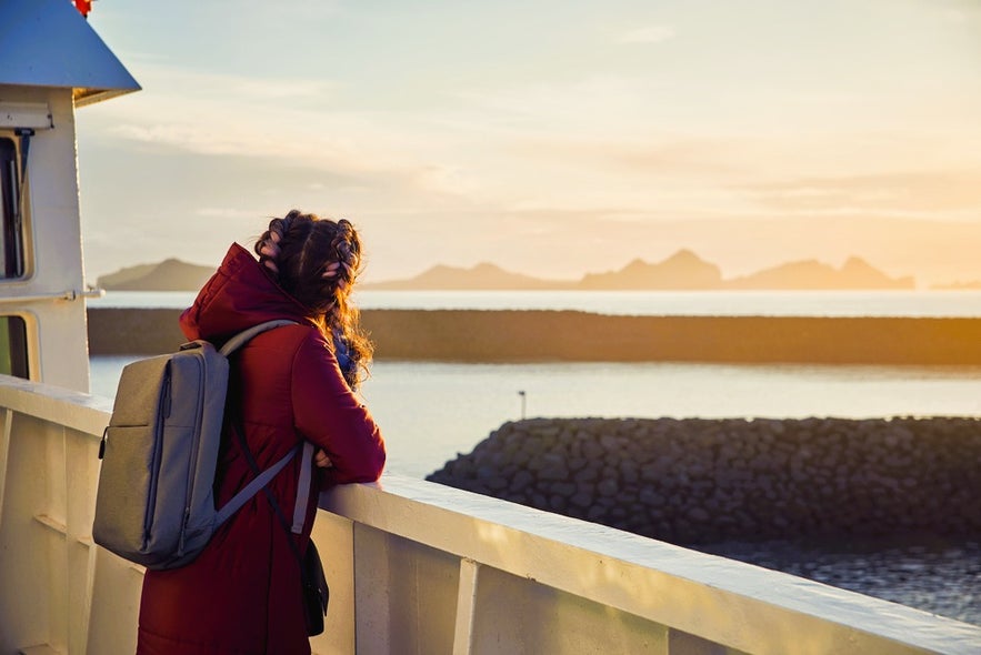 Traveler on a ferry to Iceland’s Westman Islands watching a golden sunset over Vestmannaeyjar Cliffs from the ship’s deck.