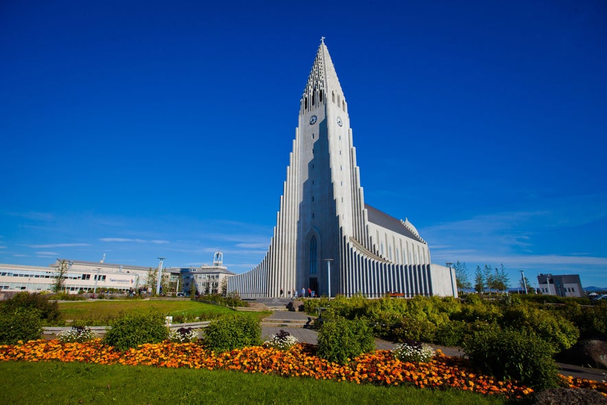 Hallgrimskirkja church under a clear blue sky in Reykjavík, Iceland, with vibrant flower beds and lush greenery in the foreground during summer.