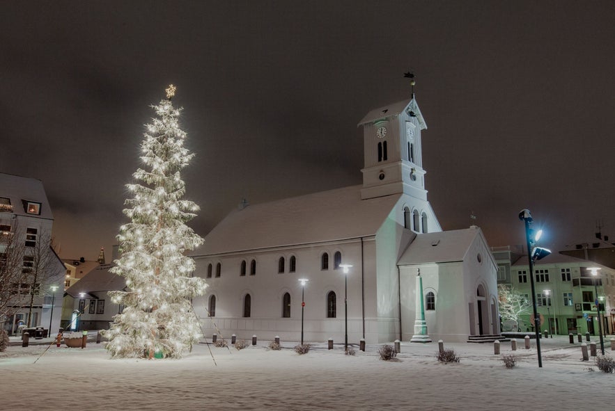 Large Christmas tree illuminated with white lights beside Reykjavík Cathedral (Dómkirkjan) in snow-covered Austurvöllur Square during winter in Iceland.