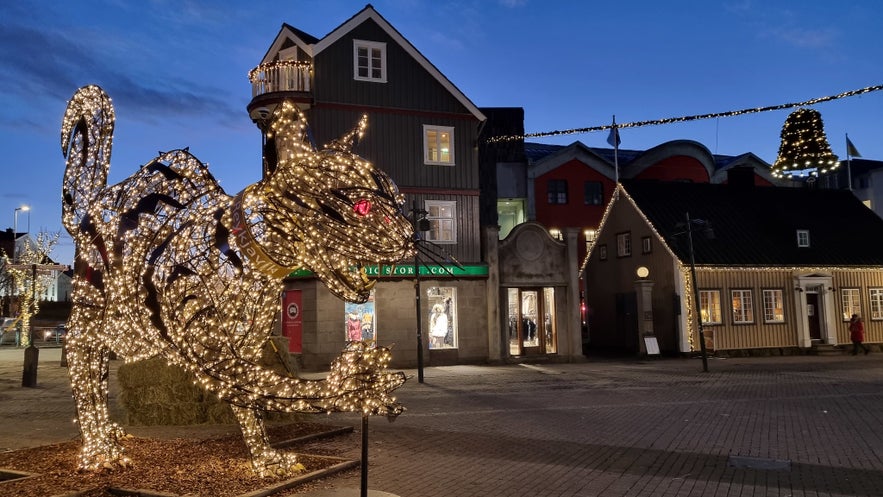 Illuminated Christmas cat sculpture with glowing red eyes in downtown Reykjavik, Iceland, surrounded by colorful buildings and festive holiday lights at dusk