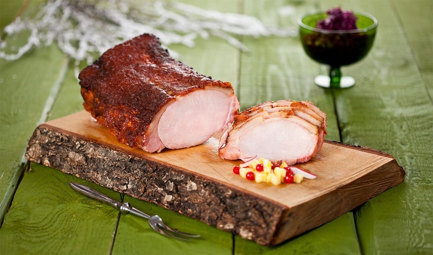 Traditional Icelandic hamborgarhryggur, a glazed and smoked pork loin roast, sliced and served on a rustic wooden board with pickled garnish, enjoyed during Iceland's Christmas dinner.