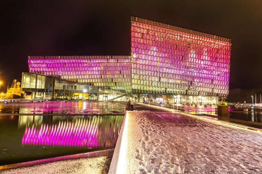 Harpa Concert Hall in Reykjavik illuminated with vibrant pink and yellow lights during winter, reflecting in surrounding water under a snowy path.