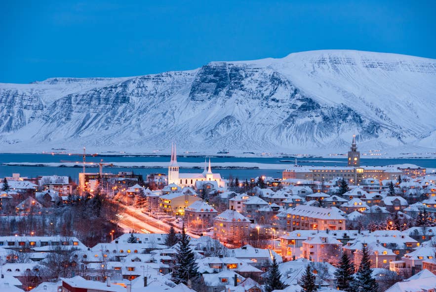 Reykjavik innevata al tramonto con le luci della città che brillano contro il Monte Esja, a testimoniare la bellezza serena dell’inverno islandese.