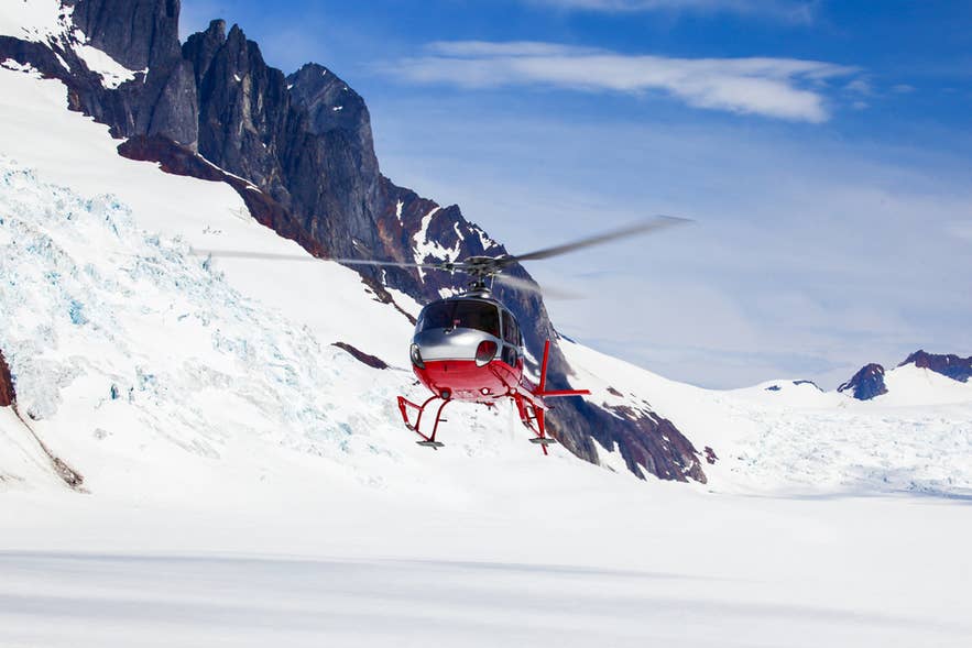 A helicopter flies low over a glacier beneath jagged peaks in Iceland.