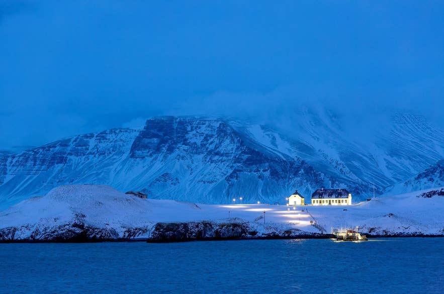 Het eiland Videy in de winterse schemering, met besneeuwde berg Esja en gloeiende lichten op de achtergrond in januari in IJsland