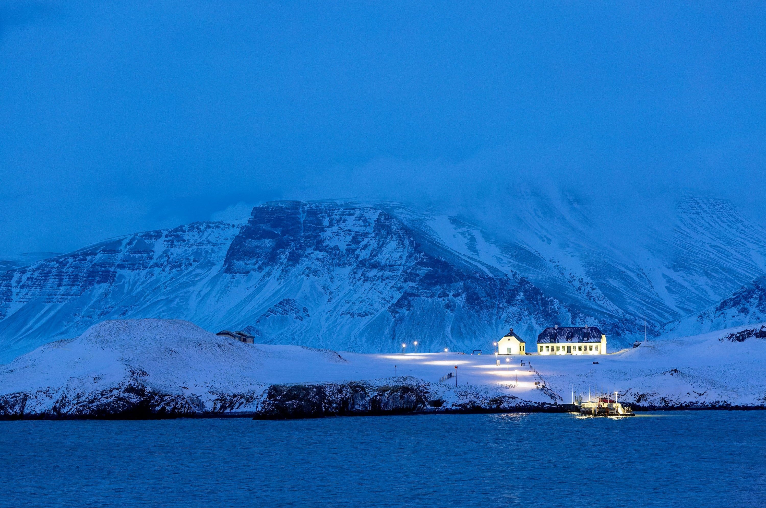 Viðey Island near Reykjavík during winter twilight, with illuminated historical buildings and snow-covered Mount Esja in the background.