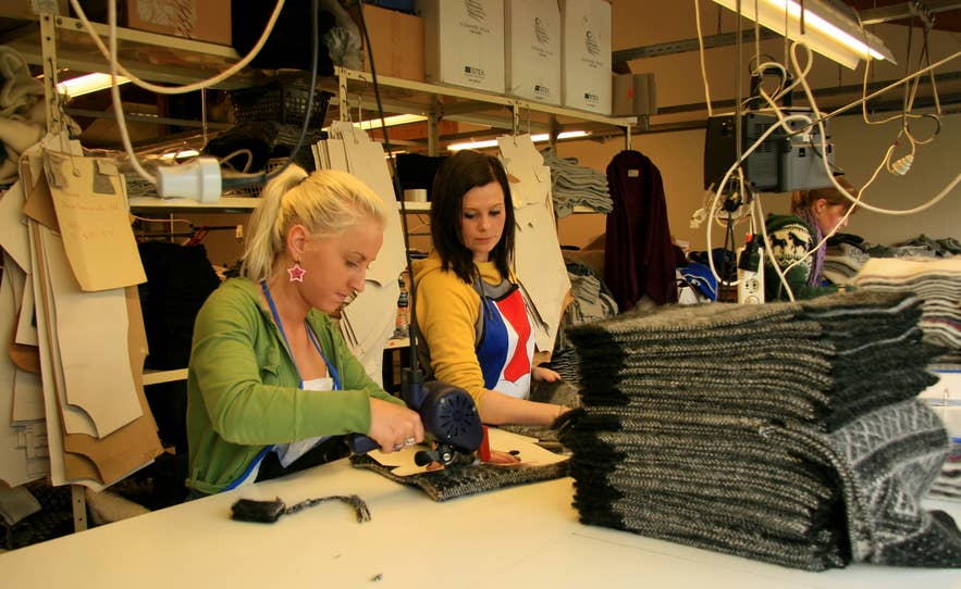 Two women working in an Icelandic knitwear workshop beside stacks of folded wool sweaters.