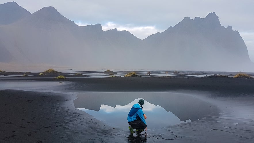 A man crouches in front of a shallow coastal pool on the black sand beach of Stokksnes Peninsula in southeast Iceland, with Vestrahorn Mountain towering in the background.