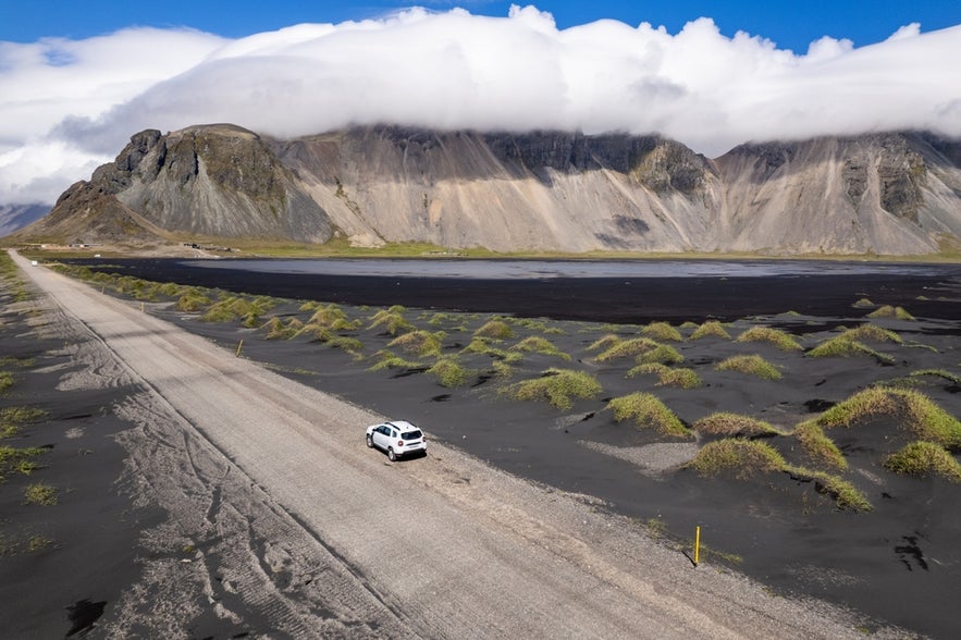 A white car travels along a sandy road on the Stokksnes Peninsula in southeast Iceland, surrounded by black sand, patches of green grass, and Vestrahorn Mountain shrouded in clouds.