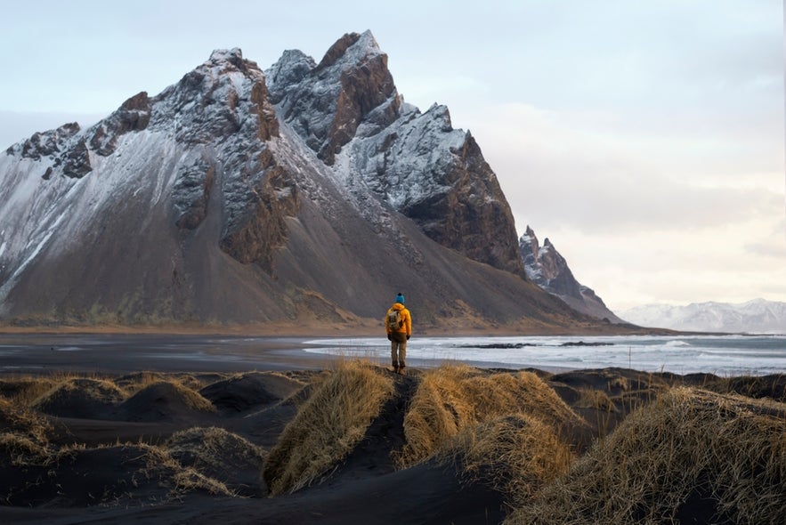 A tourist stands on the black sand beach of Stokksnes Peninsula in southeast Iceland, with Vestrahorn Mountain rising in front of him.