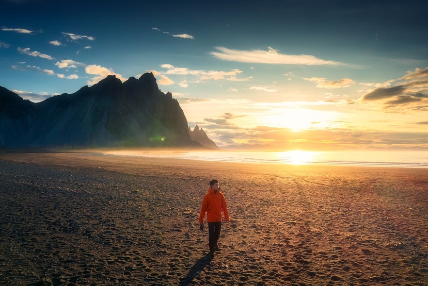 A visitor walks along the black sand beach on Stokksnes Peninsula in southeast Iceland, with the setting sun and Vestrahorn Mountain behind him.