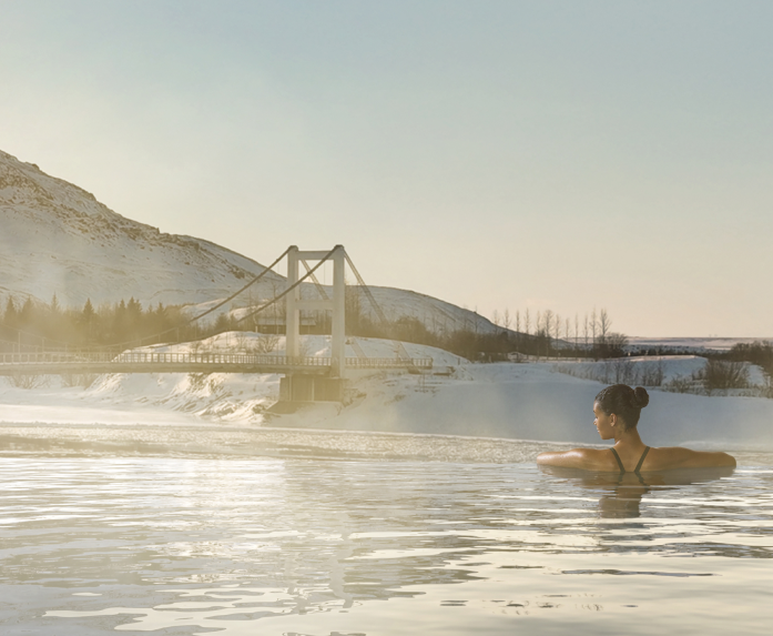 Woman relaxing in Laugaras Lagoon geothermal pool with snowy mountains and a bridge in South Iceland.