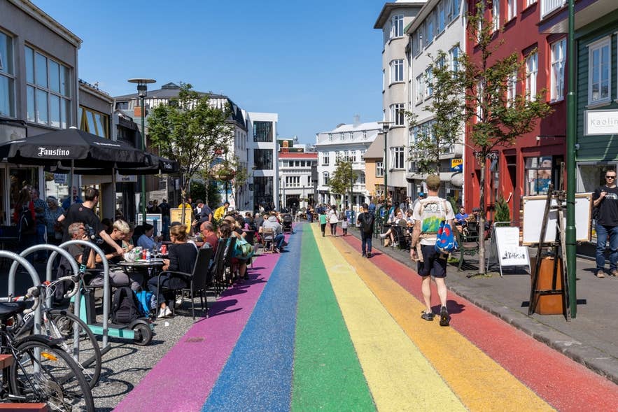Rainbow-painted Skolavordustigur Street in downtown Reykjavik with outdoor cafés, pedestrians, and colorful city buildings on a sunny day.