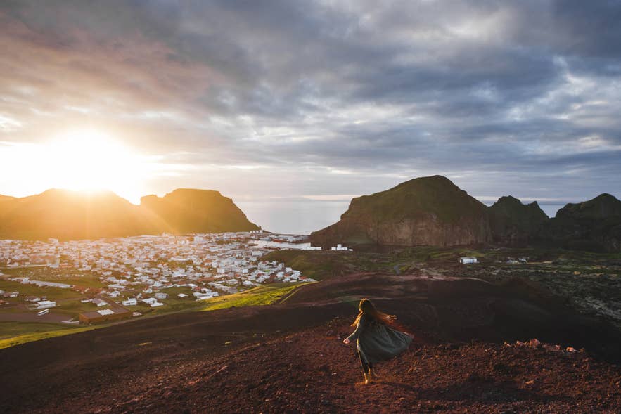 En ung, glad kvinne nyter solnedgangen på toppen av Eldfell-vulkanen, med panoramautsikt over Vestmannaeyjar, Island.