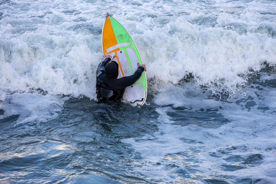 Surfer in Iceland swimming with a surfboard at a black sand beach.