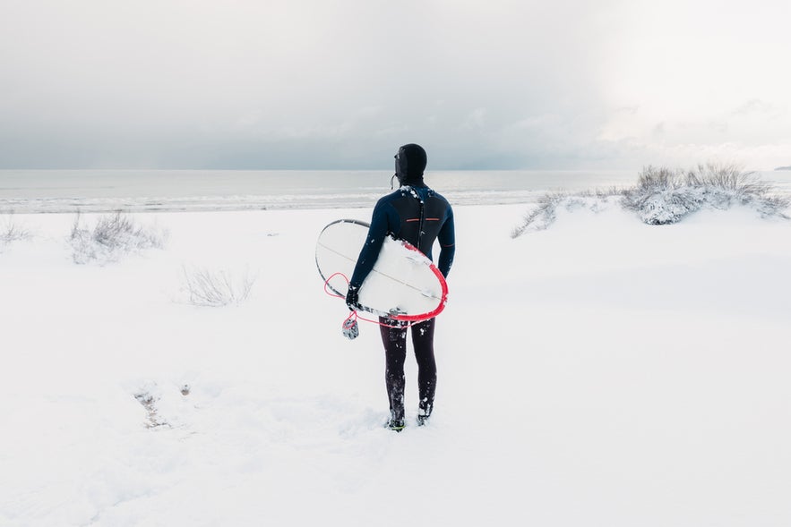 Surfing in Iceland in winter, with a surfer in a wetsuit carrying a surfboard on a snowy beach.