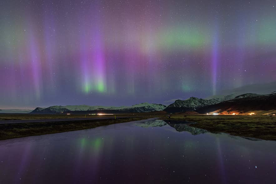 Paars en groen noorderlicht dat weerspiegelt in het water, met besneeuwde bergen en een klein IJslands dorp op de achtergrond.