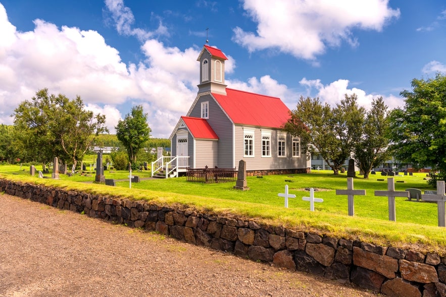 A quaint church with a red roof and surrounding cemetery in Reykholt, Iceland, under a bright blue sky with scattered clouds.