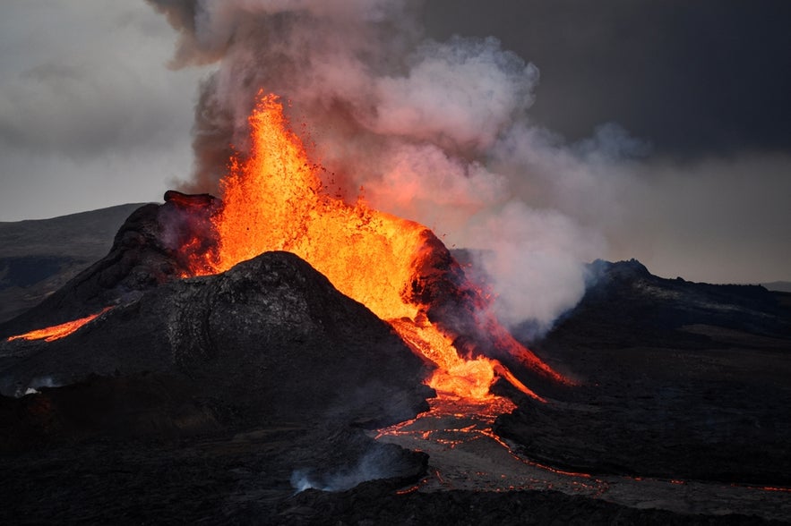 雷克雅内斯半岛法格拉达尔火山喷发，熔岩从火山口喷涌而出，顺着漆黑的火山地形不断流淌。