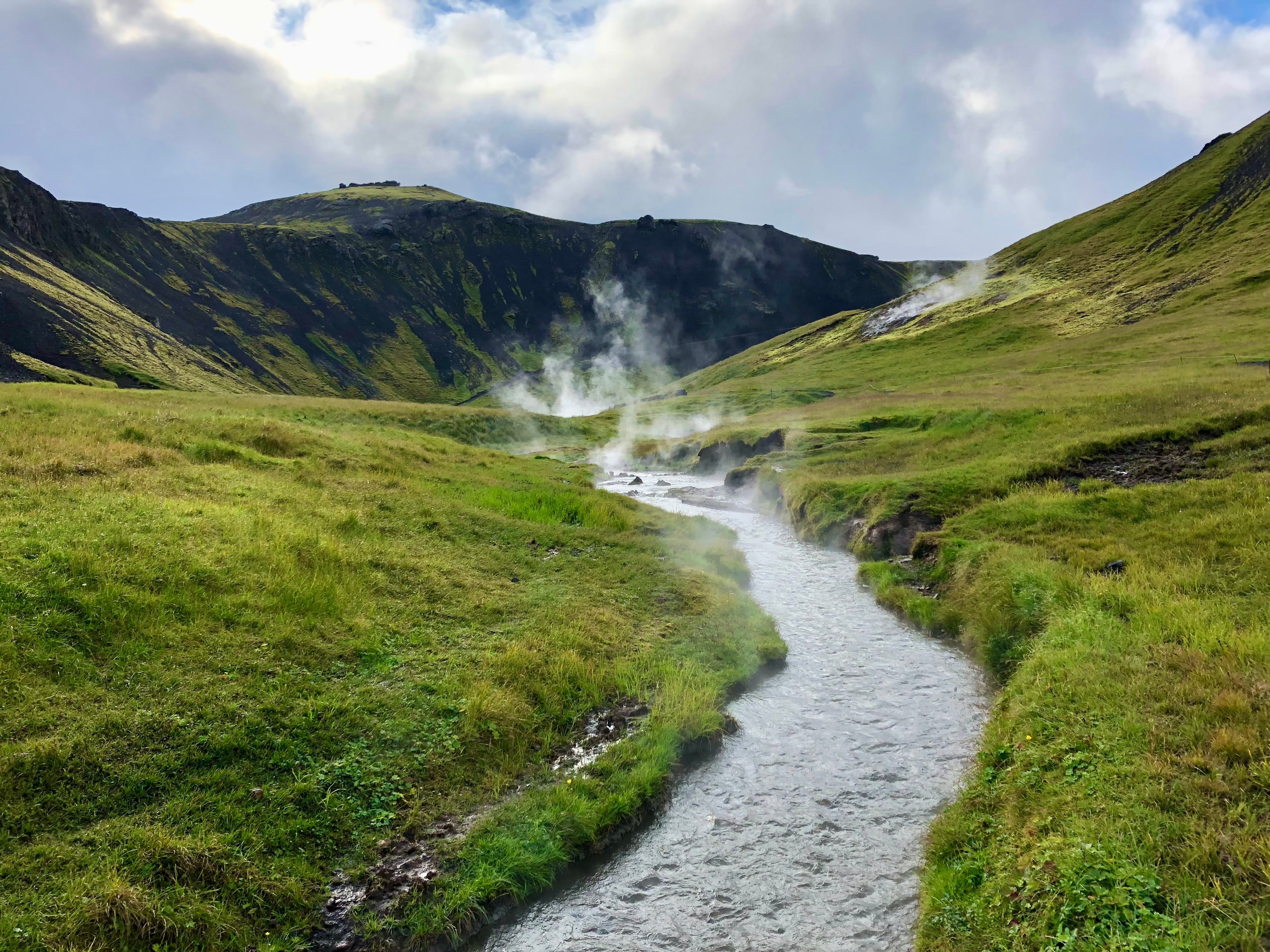 Steaming Reyjaladur River flows through a lush green valley, leaving trails of steam on its path.