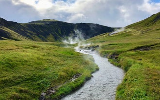 Steaming Reyjaladur River flows through a lush green valley, leaving trails of steam on its path.