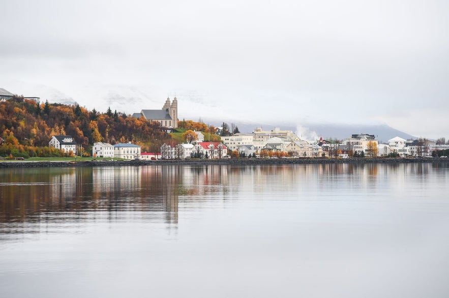utumn view of Akureyri in North Iceland with colorful foliage, calm fjord reflections, and the prominent Akureyrarkirkja church on a hill under overcast skies.