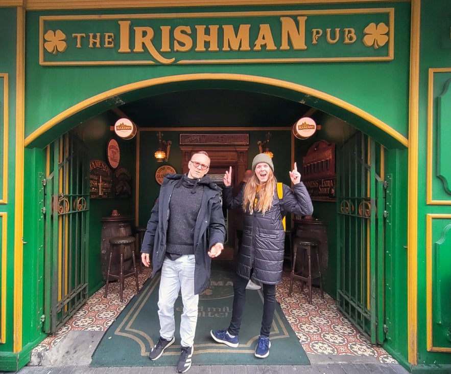 Entrance of The Irishman Pub in Reykjavik with two happy visitors posing under the green and gold sign.