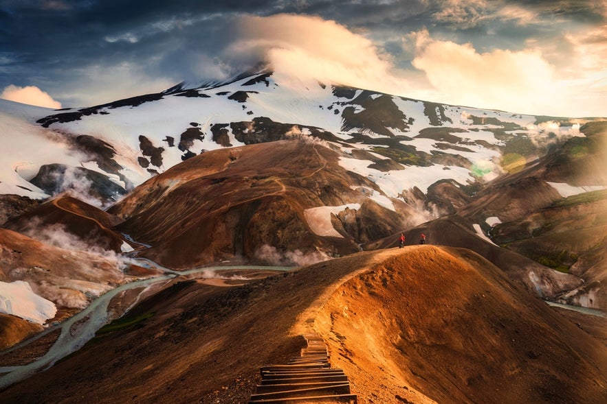 A wooden path in Kerlingarfjoll, with geothermal vents, volcanic hills, and snow-capped mountains in the background.