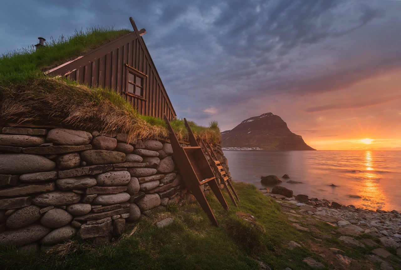 A turf-roofed hhut sits on an elevated rocky spot overlooking a fjord with views of distant mountains.