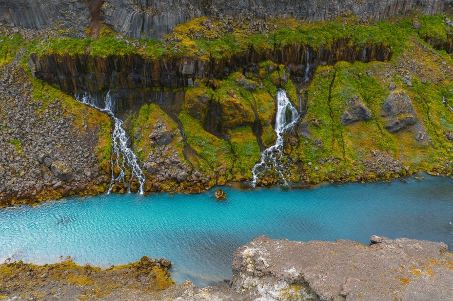 Turquoise water flows through Sigoldugljufur Canyon, Iceland, with moss covered cliffs and cascading waterfalls over basalt rock formations.
