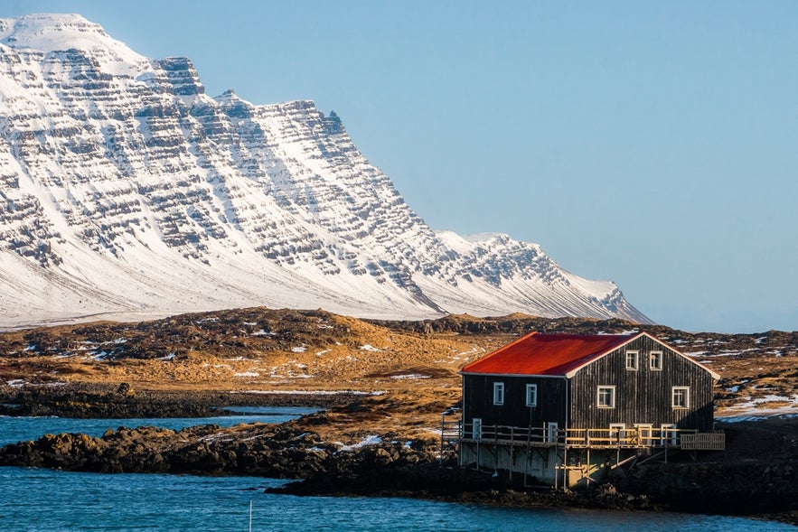 Beautiful winter view of a traditional black house by the shore in Djúpivogur, East Iceland, with snowy mountains in the background.