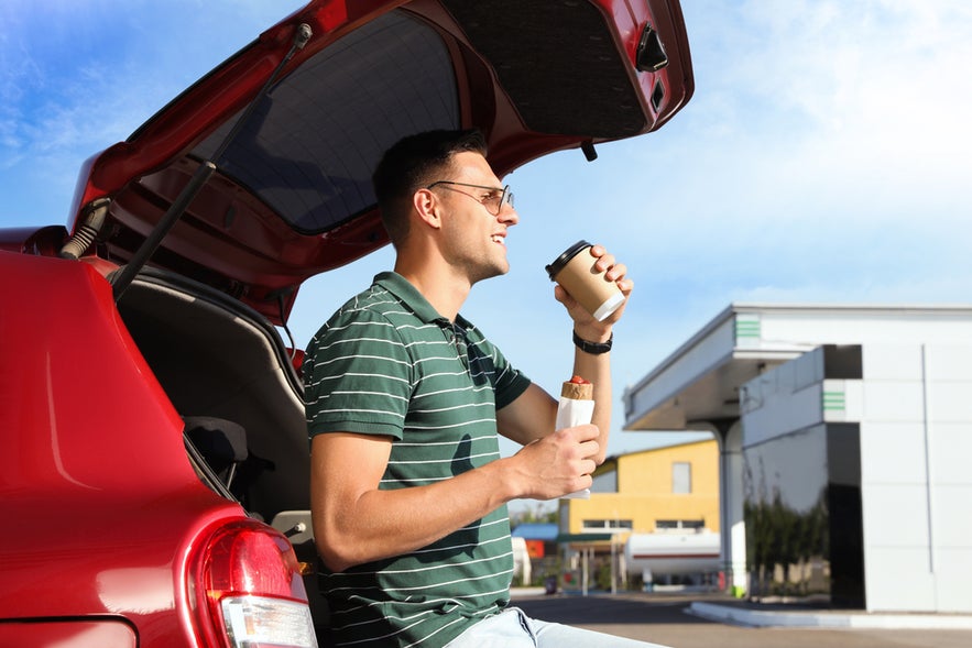 Young man with hot dog drinking coffee near car at gas station in Iceland.