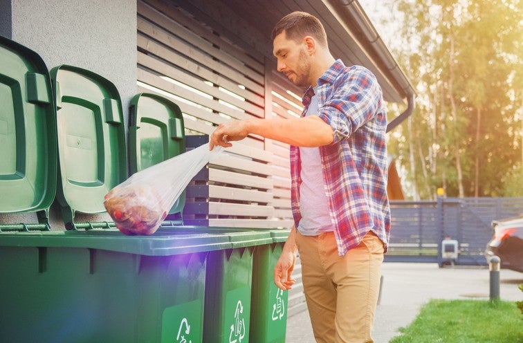A man throwing away two plastic bags of trash into a bin outdoors.