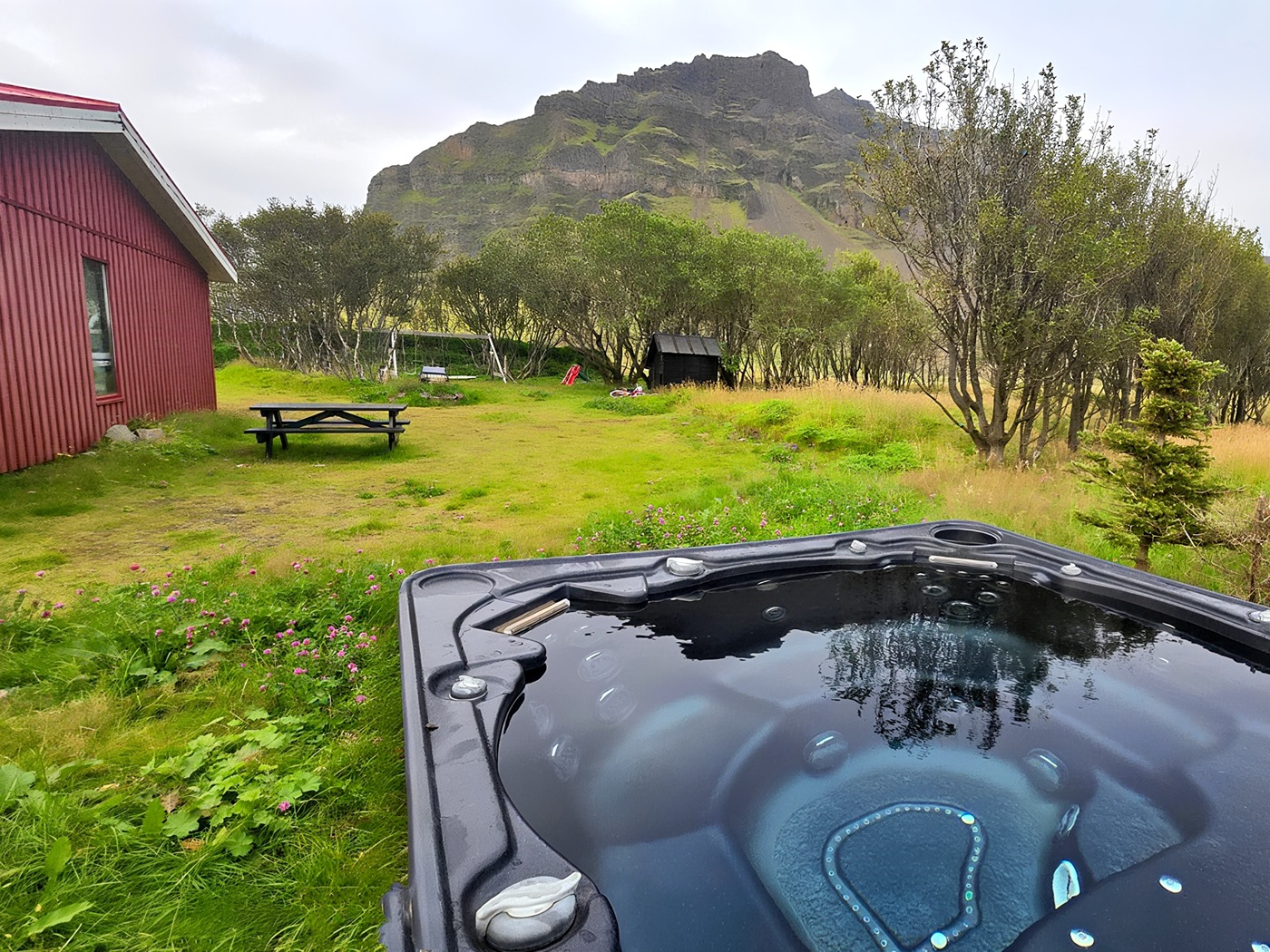 Utsikt fra boblebadet over et jorde med trær og fjell i det fjerne, hytta delvis synlig til venstre.