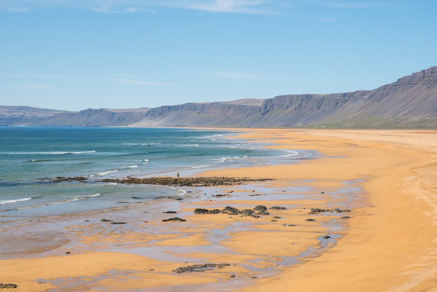 Ampia spiaggia dorata di Raudasandur nei Westfjords islandesi, con onde leggere sulla riva e montagne ripide sullo sfondo sotto un cielo azzurro.