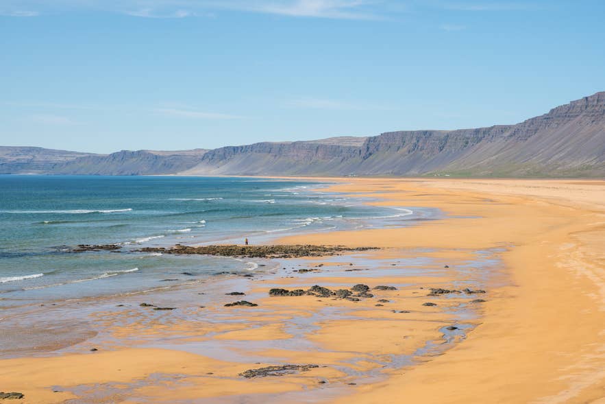 Brede goudkleurige zandstrand bij Raudasandur in de Westfjorden van IJsland, met zachte golven die de kust raken en steile bergen op de achtergrond onder een heldere blauwe lucht.