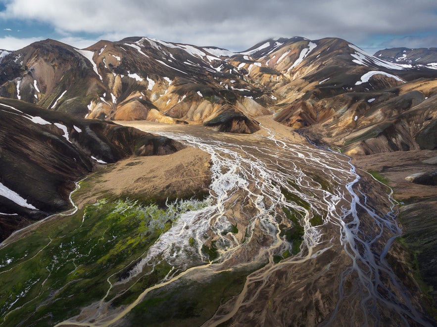 Aerial view of Landmannalaugar in Iceland’s Highlands, showing colorful rhyolite mountains, braided glacial rivers, green moss, and patches of snow under a cloudy sky.
