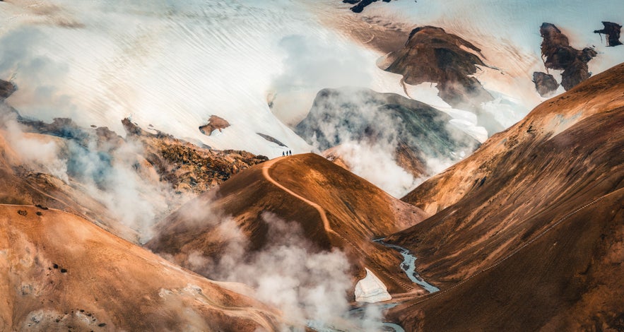 Hikers walking along a winding trail through the steaming rhyolite mountains of Hveradalir in Kerlingarfj&ouml;ll, Iceland, with patches of snow and geothermal steam rising from the colorful slopes.