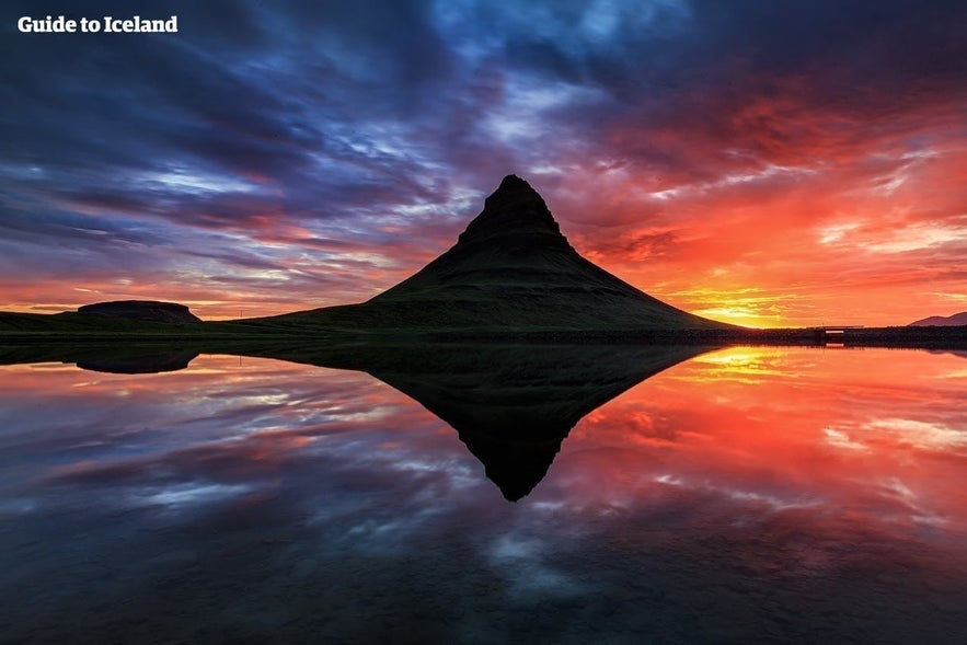 Monte Kirkjufell al sole di mezzanotte con colori vividi del tramonto e riflesso perfetto nell’acqua calma.