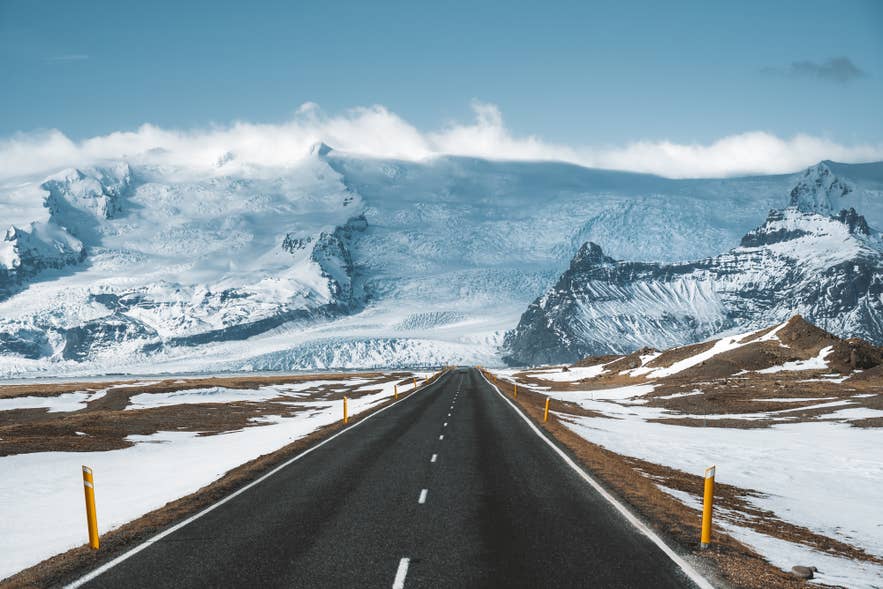 Straight road leading toward the snow-covered mountains and glaciers of Vatnajökull National Park in Iceland, with patches of snow along the roadside under a clear blue sky.