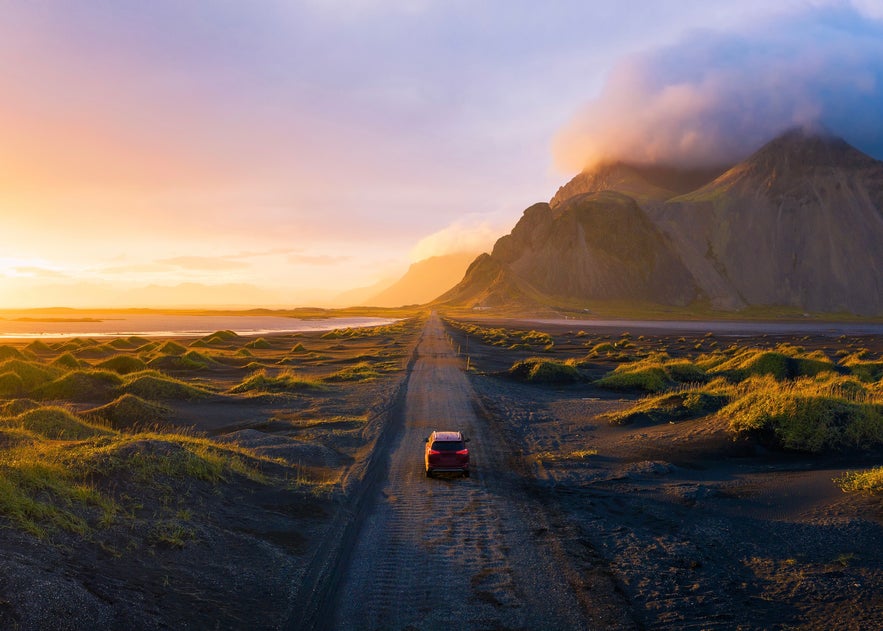 Red car driving on a black sand road at Stokksnes near Vestrahorn mountain in Iceland, with grassy dunes and a glowing sunset sky.