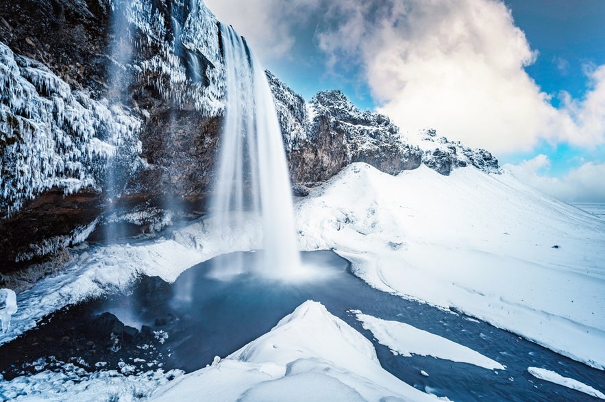 Seljalandsfoss Waterfall in South Iceland is partly frozen in winter, with snow-covered cliffs and icy surroundings.