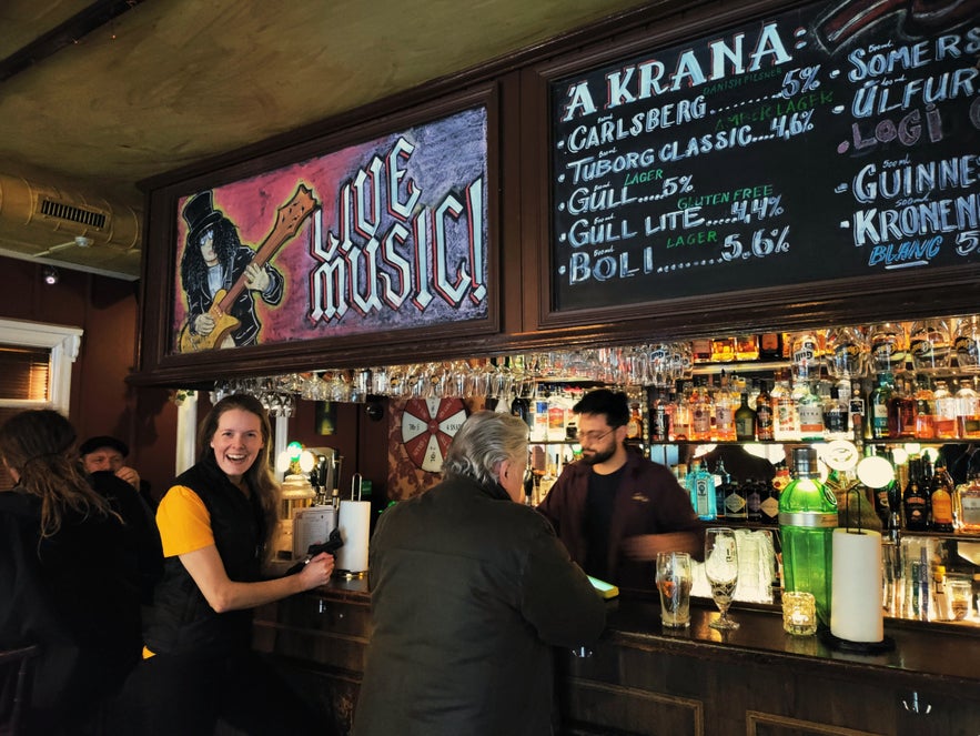 Live music sign and busy bar counter at Den Danske Kro in Reykjavík, with smiling staff and customers.
